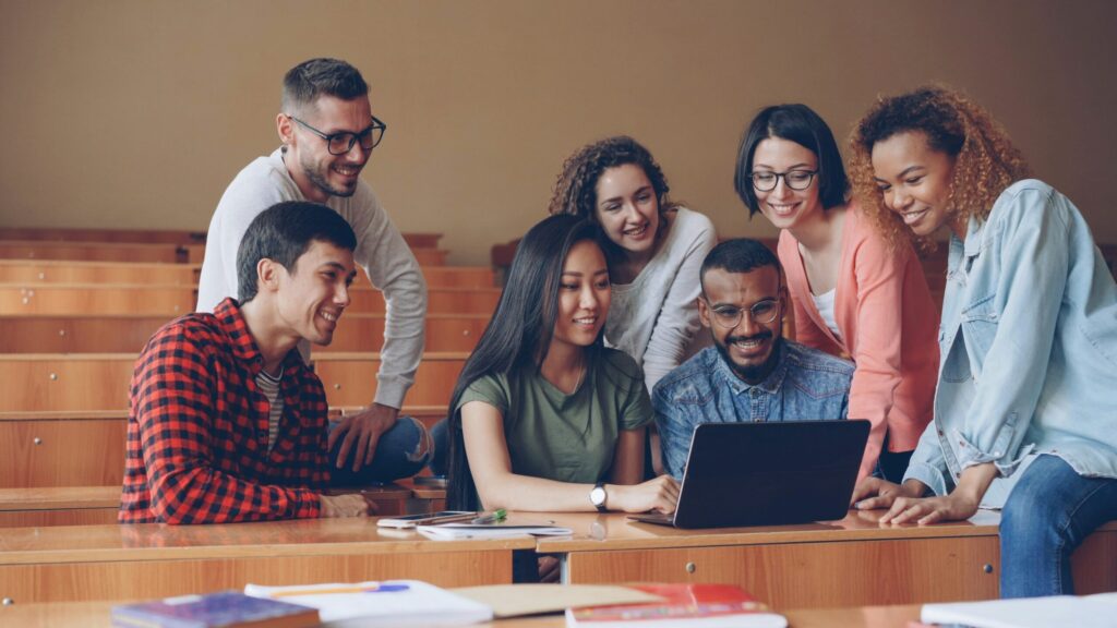 Six university students collaborate around a laptop in a lecture hall, smiling and discussing their work together.