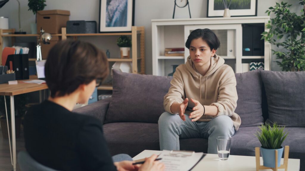 A young person sits on a sofa speaking with a counselor, who is seated opposite and taking notes. The calm, home-like room features bookshelves, plants, and a coffee table with a glass of water.